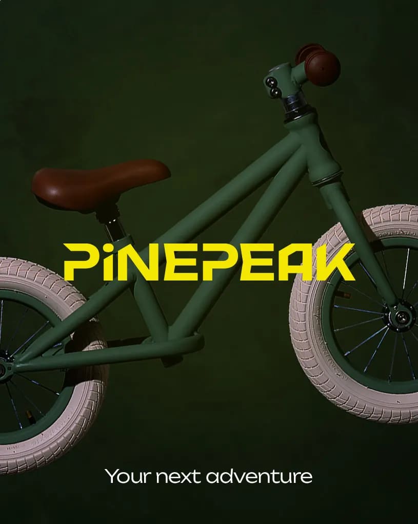 Mørkegrøn løbecykel til børn med brede lyse dæk og brun sadel. Gul tekst: PINEPEAK. Hvid tekst: Your next adventure.